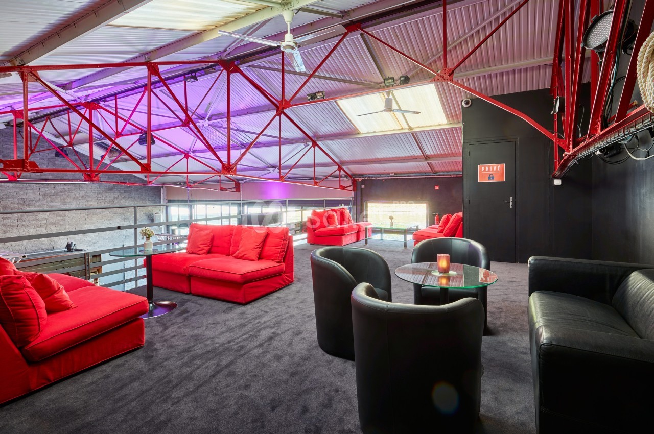 Salon industriel avec canapés rouges, fauteuils noirs et tables basses, situé sous une structure métallique rouge.