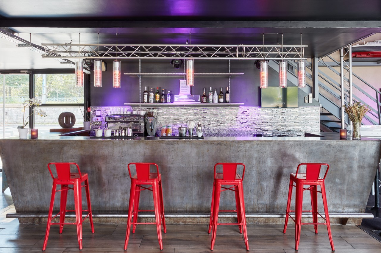 Bar moderne avec comptoir métallique, tabourets rouges, étagères de bouteilles et éclairage violet.