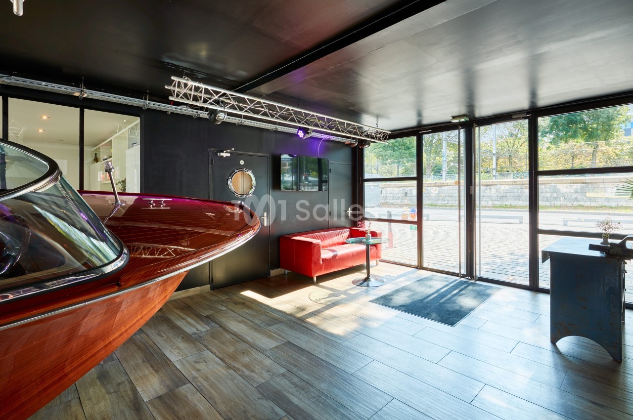 Intérieur moderne avec un bateau en bois, canapé rouge, grandes baies vitrées et vue sur un espace extérieur arboré.