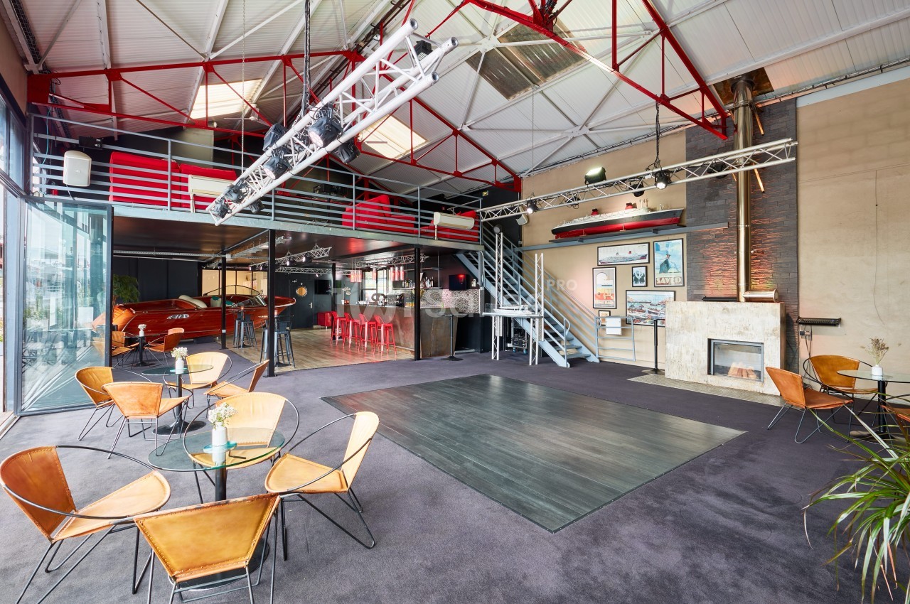 Salle spacieuse avec mezzanine, tables en verre, chaises design, bar rouge et grande baie vitrée laissant entrer la lumière.