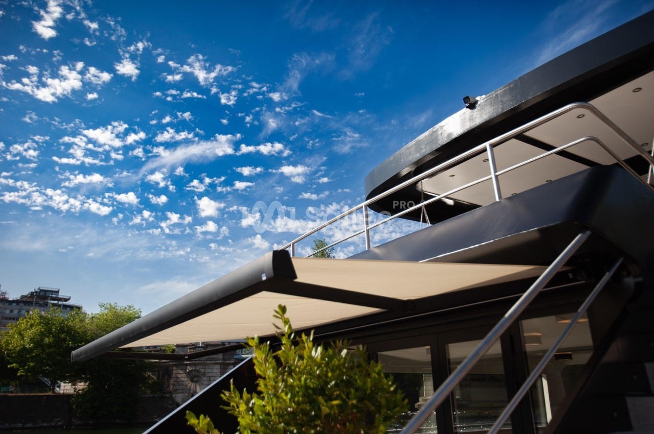 Vue en contre-plongée d'une terrasse de bateau moderne avec un store déployé, sous un ciel bleu parsemé de nuages.