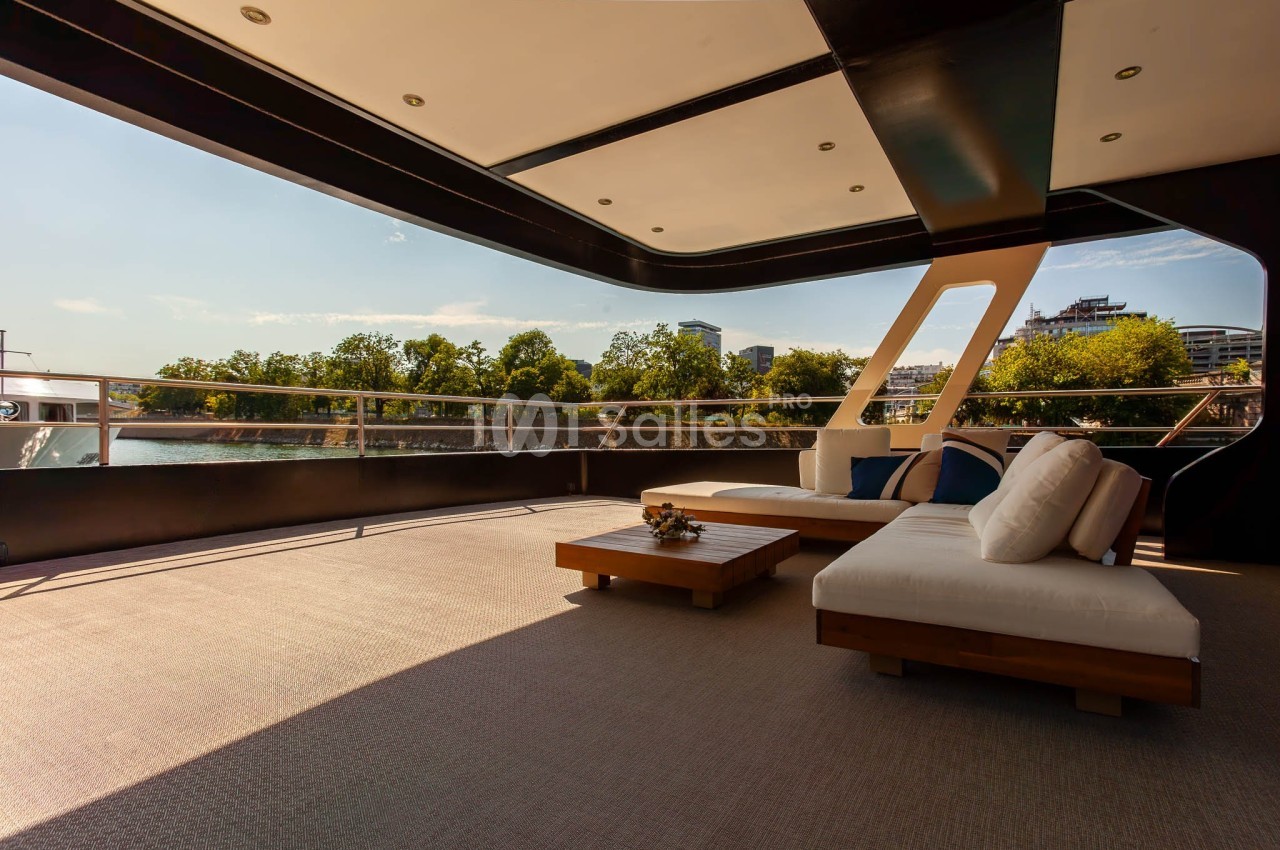 Terrasse d’un yacht avec mobilier en bois et coussins, offrant une vue dégagée sur des arbres et un plan d’eau.