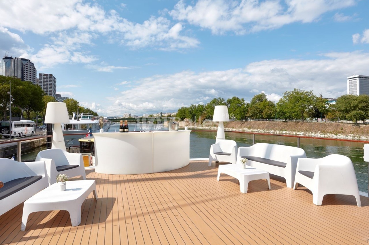 Terrasse d'un bateau avec mobilier moderne, vue sur un fleuve bordé d'arbres et bâtiments en arrière-plan.
