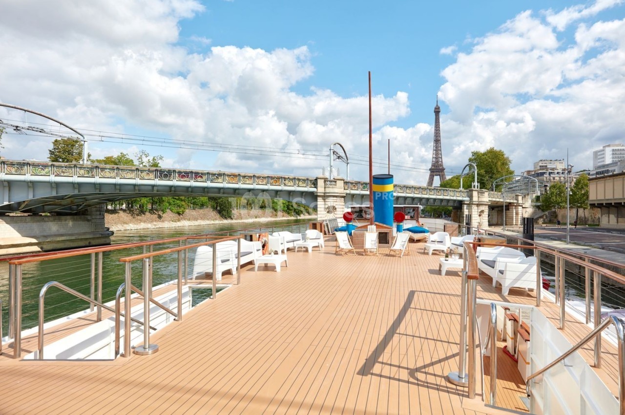 Pont supérieur d'un bateau avec des chaises longues, vue sur la Seine, un pont ferroviaire et la tour Eiffel en arrière-plan.