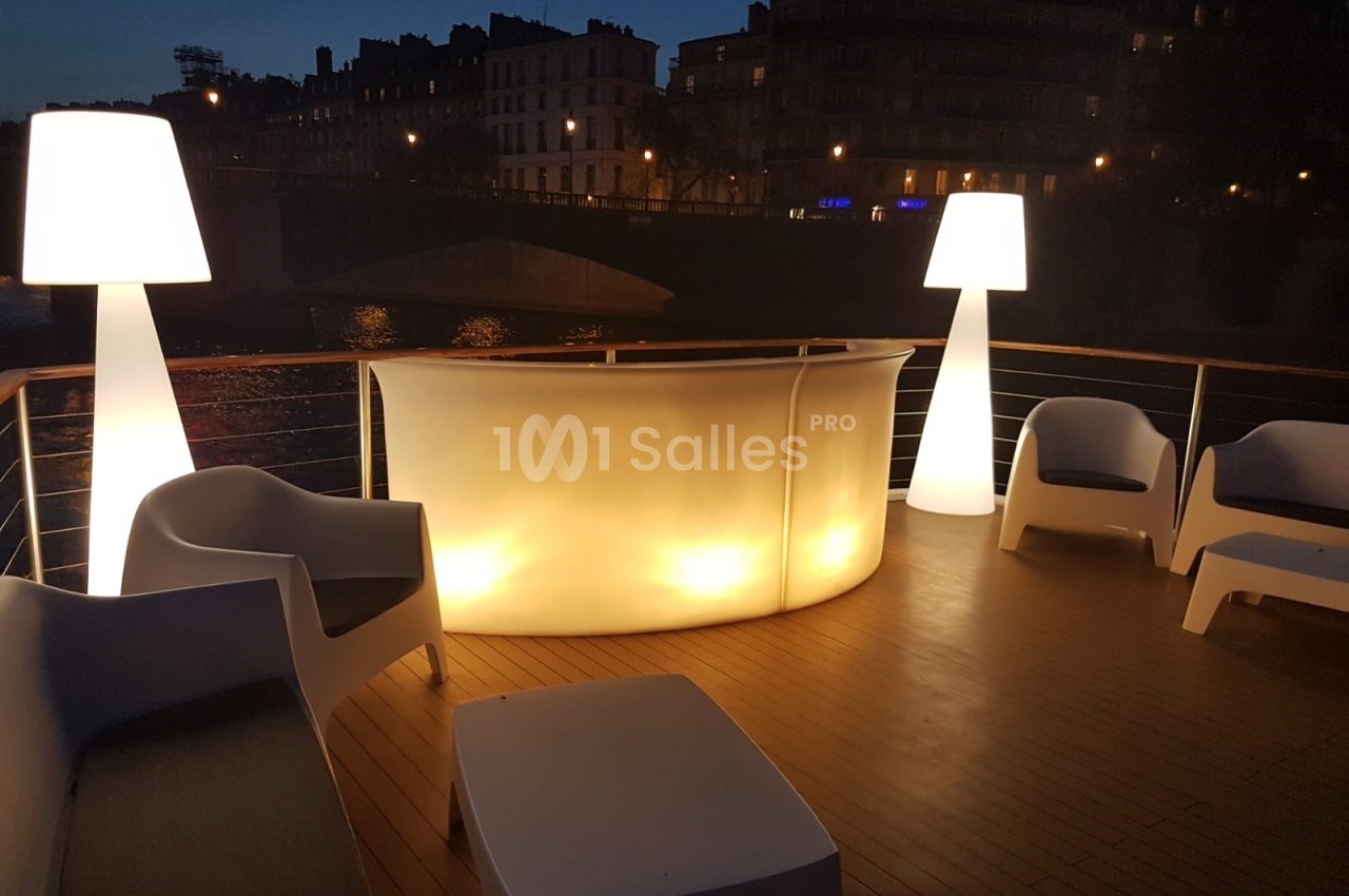 Terrasse en bois éclairée de nuit avec un bar lumineux, des fauteuils blancs et des lampes design près d'une rivière.