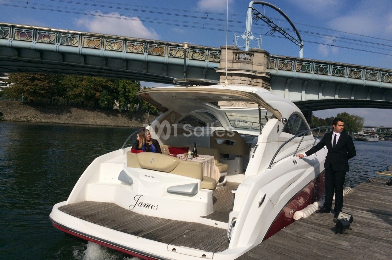 Un yacht amarré près d'un quai sous un pont, avec une femme assise et un homme debout sur le ponton.