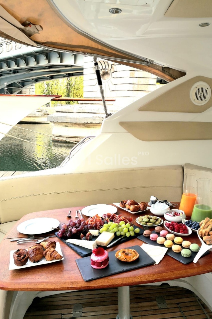Petit-déjeuner composé de viennoiseries, fruits, macarons et jus, servi sur une table en bois à bord d'un bateau.