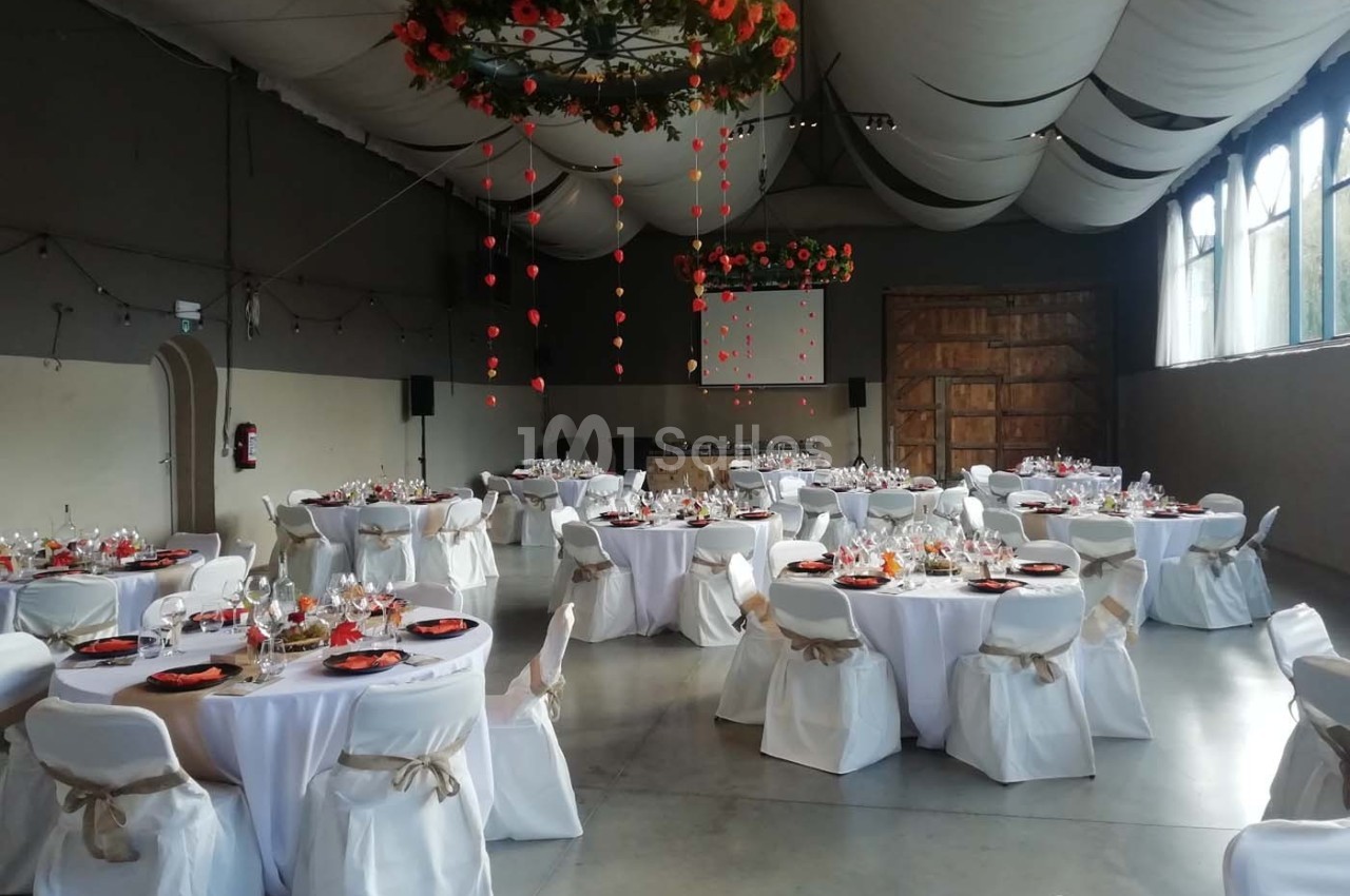 Salle de réception décorée avec des tables rondes dressées, nappes blanches et guirlandes suspendues au plafond.