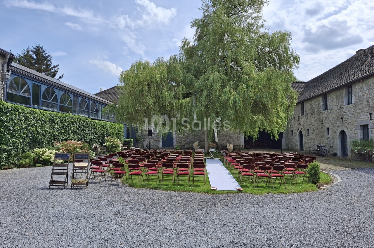 Chaises rouges disposées en extérieur sous un grand arbre pour une cérémonie, entourées de bâtiments en pierre.