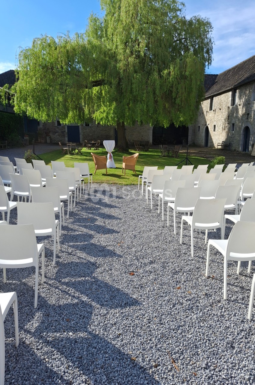 Chaises blanches disposées en rangées face à un arbre et une table décorée dans une cour extérieure ensoleillée.