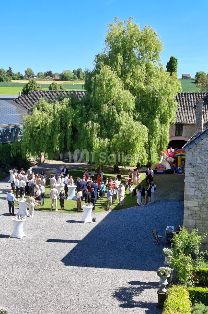 Groupe de personnes rassemblées dans une cour extérieure ensoleillée, entourée de bâtiments en pierre et de verdure.