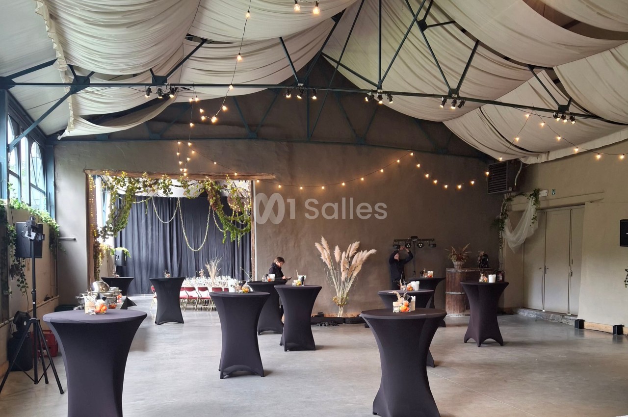 Salle de réception décorée avec des guirlandes lumineuses, des tables hautes noires et des éléments végétaux.