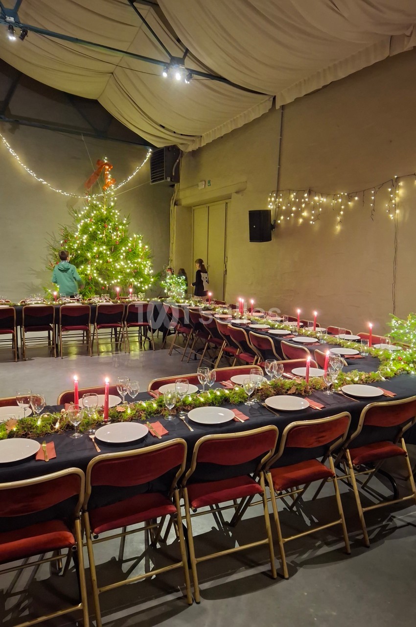 Salle décorée pour un repas festif avec longues tables dressées, guirlandes lumineuses et grand sapin illuminé.