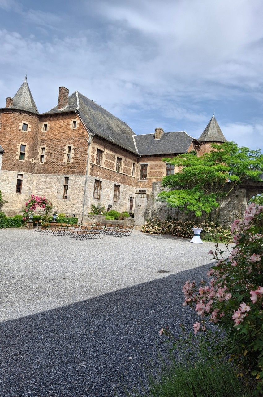Cour d’un château en pierre et briques rouges, avec tours, fleurs, mobilier extérieur et ciel partiellement nuageux.
