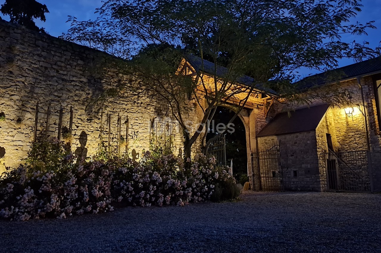 Entrée en pierre éclairée de nuit, entourée de fleurs et d'arbres, avec un portail en fer forgé.