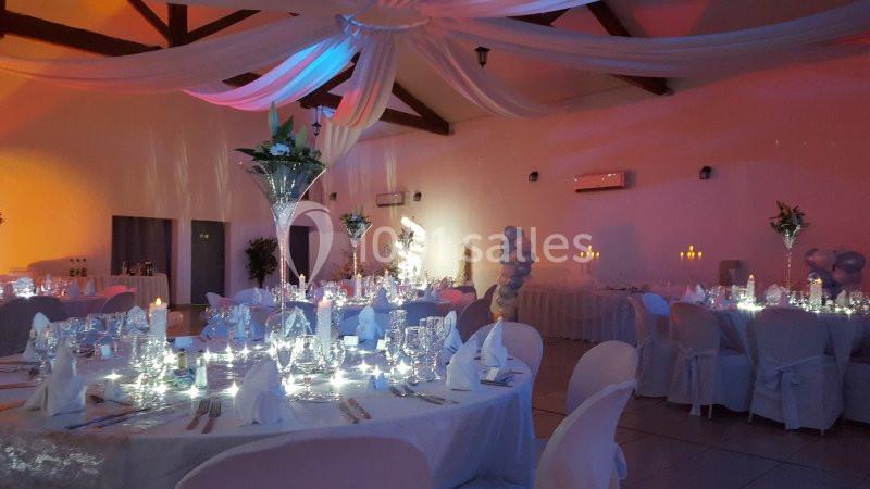 Salle de réception décorée avec des tables rondes dressées, nappes blanches, éclairage tamisé et drapés suspendus.