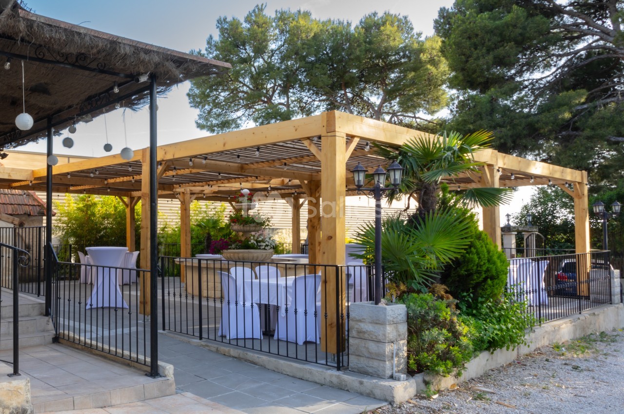 Pergola en bois avec tables dressées et chaises blanches, entourée de végétation et éclairée par des lampes suspendues.