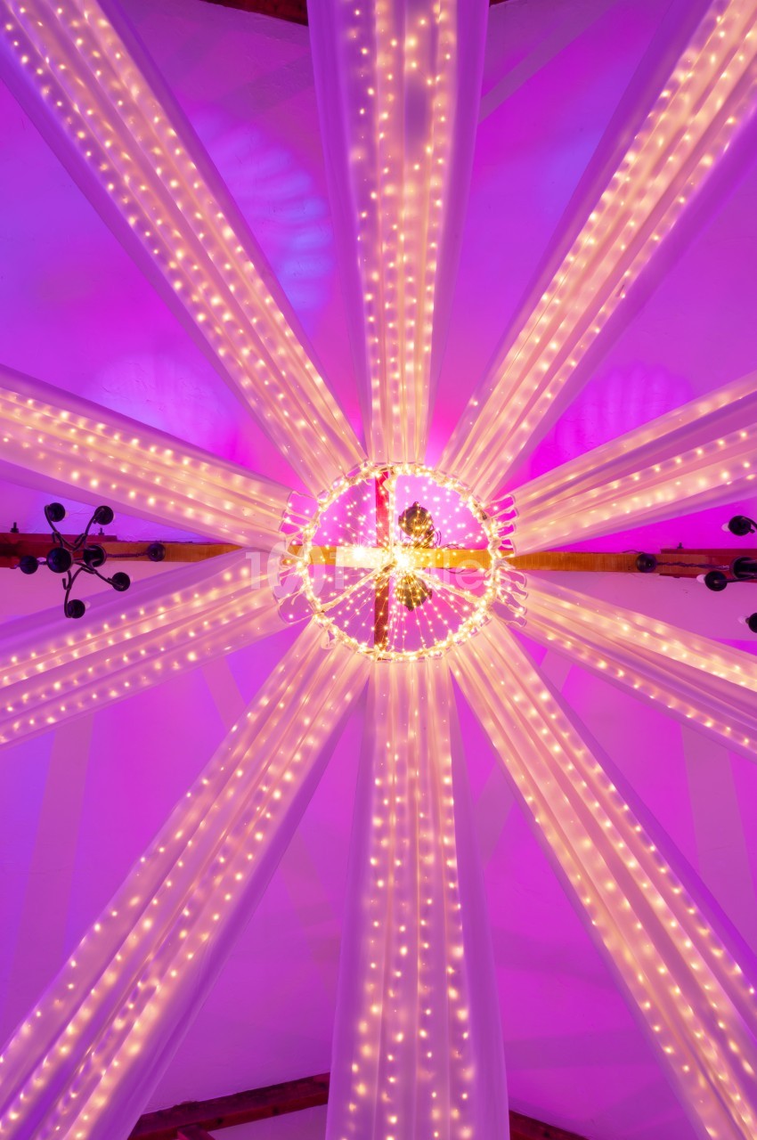 Structure lumineuse circulaire suspendue, ornée de guirlandes LED et de drapés blancs, éclairée en rose.
