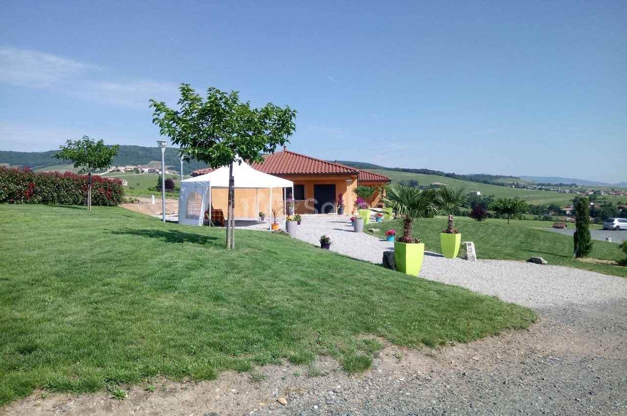 Maison avec toit en tuiles, terrasse, tente blanche et jardin verdoyant dans un paysage rural ensoleillé.