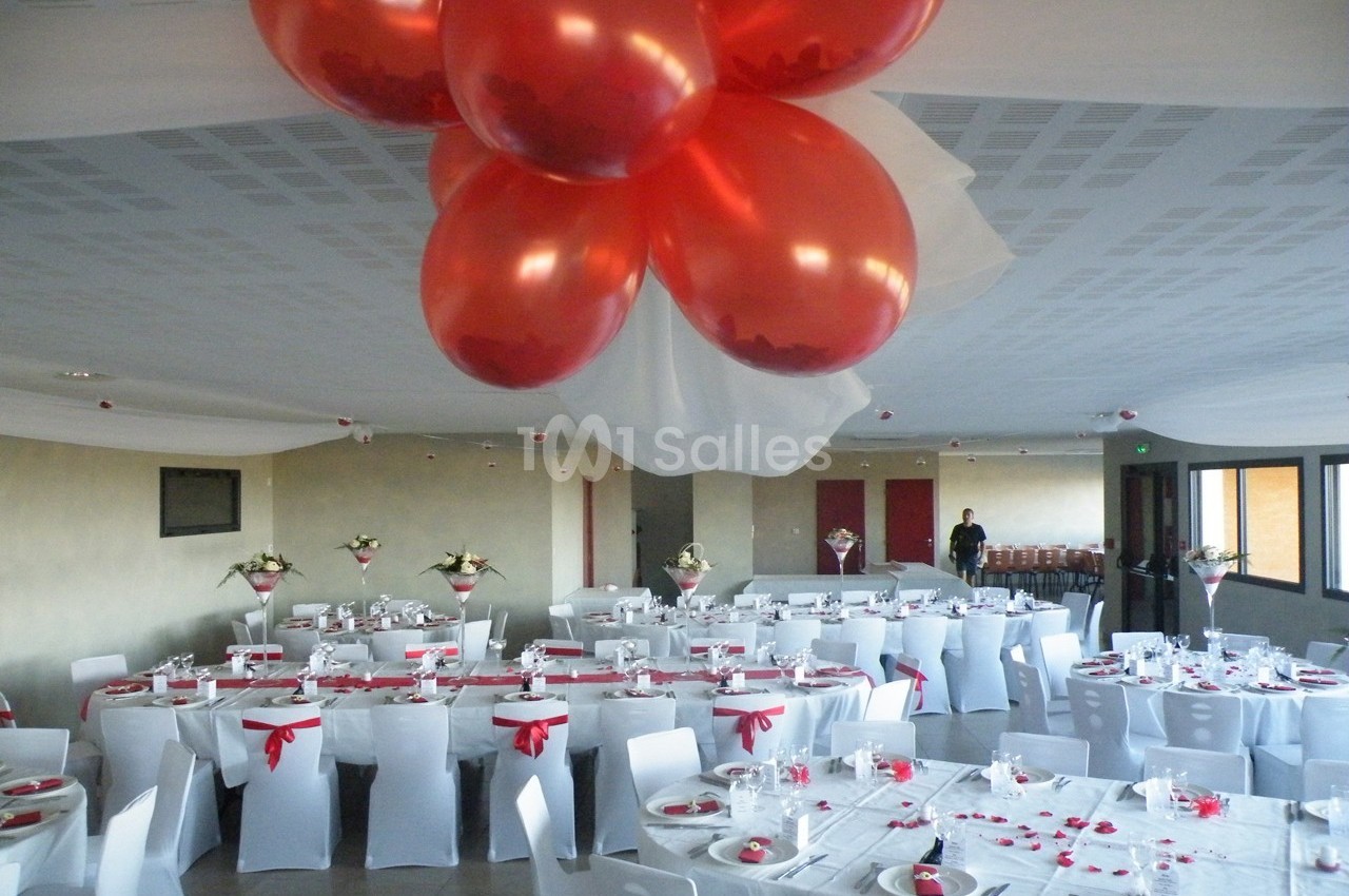 Salle de réception décorée avec des ballons rouges au plafond, tables dressées avec nappes blanches et accents rouges.