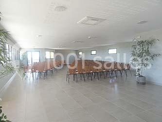 Salle lumineuse avec rangées de chaises alignées face à une zone dégagée, entourée de plantes et de fenêtres.