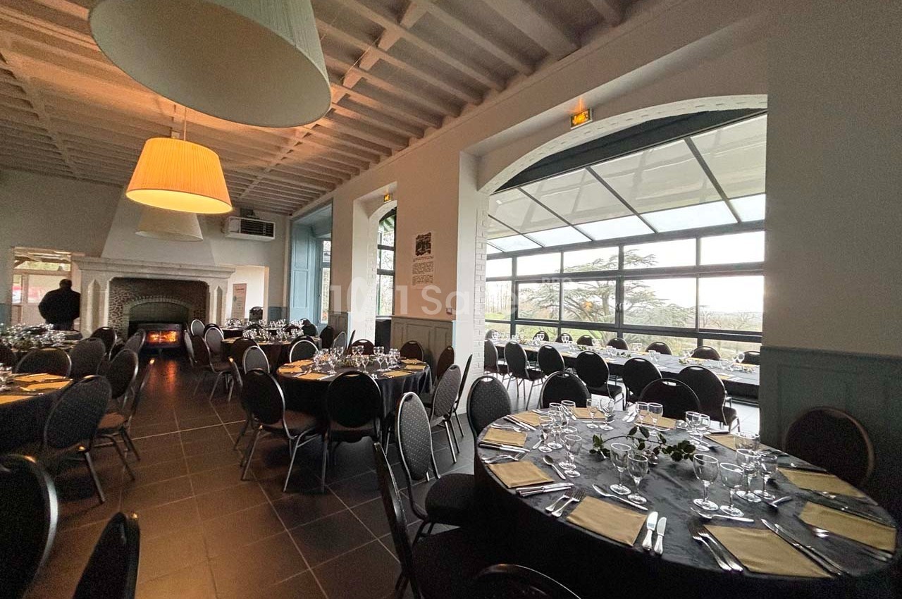 Salle de réception lumineuse avec tables rondes dressées, grandes fenêtres et cheminée en arrière-plan.