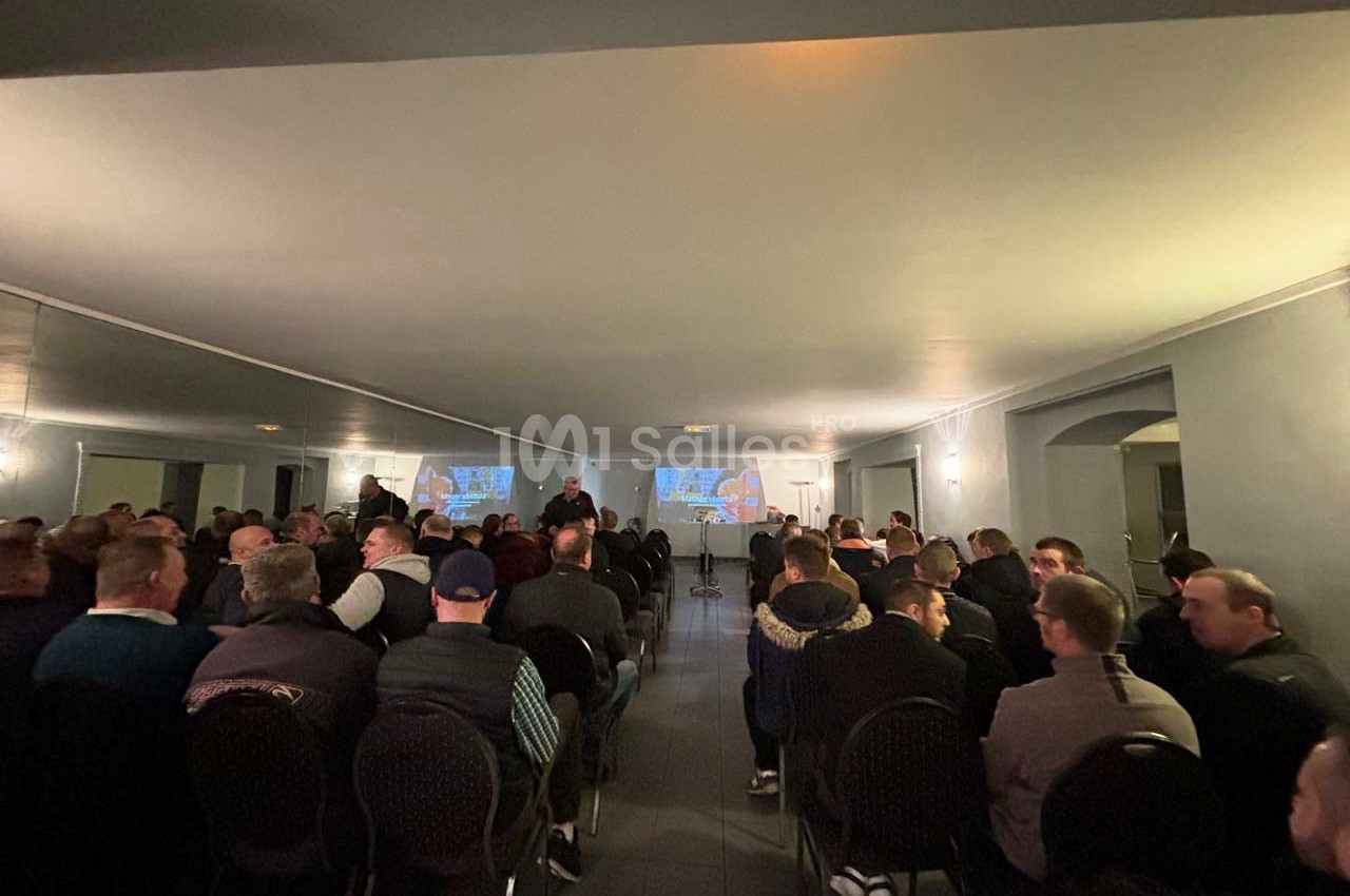 Salle remplie de personnes assises, regardant une présentation projetée sur deux écrans au fond de la pièce.