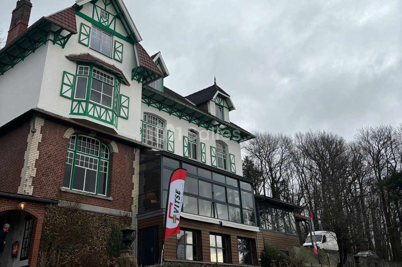 Façade d'une grande maison à colombages verts avec une extension vitrée, entourée d'arbres sous un ciel nuageux.