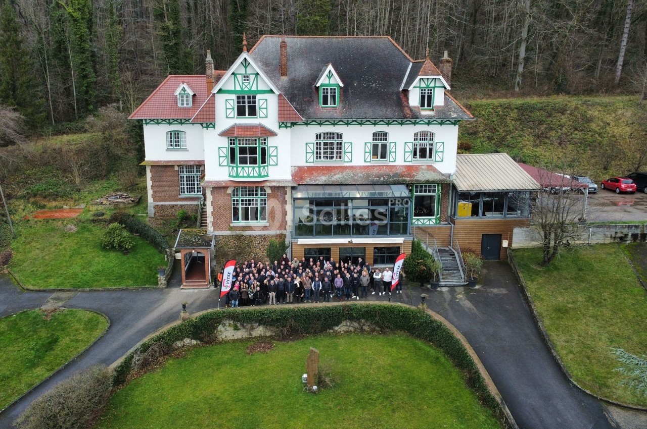 Photo aérienne d'un grand bâtiment de style traditionnel entouré de verdure, avec un groupe de personnes rassemblées devant.