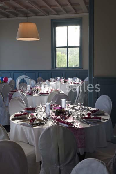 Salle de réception avec tables rondes dressées, nappes blanches, vaisselle élégante et lumière naturelle.