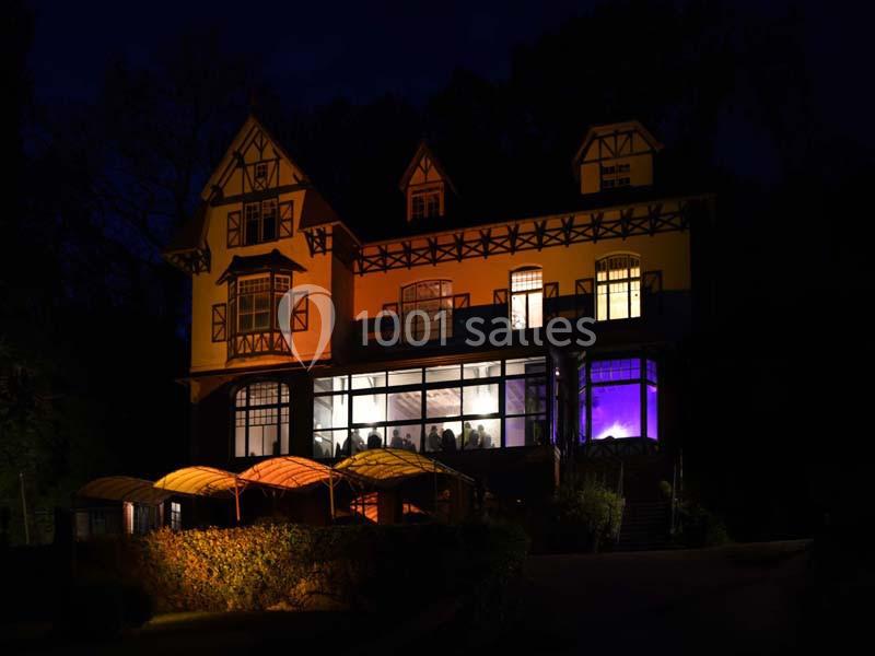 Façade d'une grande maison à colombages éclairée la nuit, avec des fenêtres illuminées et un jardin sombre au premier plan.