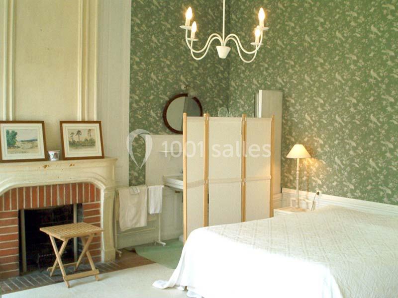 Chambre avec lit double, cheminée, paravent devant un lavabo, mur vert à motif floral et lustre suspendu.