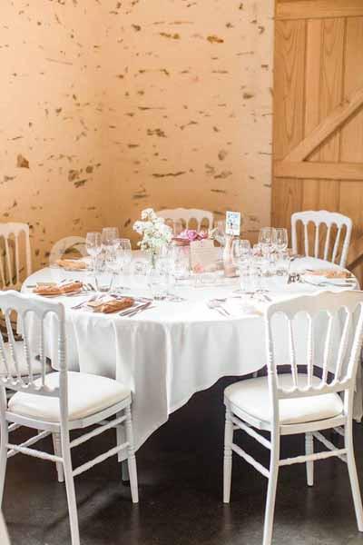 Table ronde élégamment dressée avec nappes blanches, chaises blanches et décoration florale dans une salle aux murs clairs.