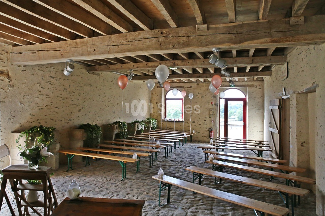 Salle rustique avec murs en pierre, poutres apparentes, bancs en bois, ballons suspendus et lumière naturelle.