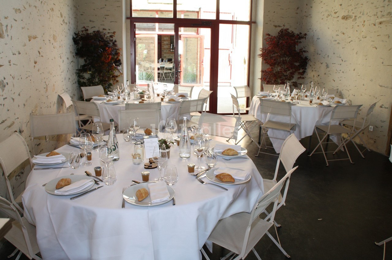 Salle de réception avec tables rondes dressées, nappes blanches, vaisselle élégante et chaises pliantes.