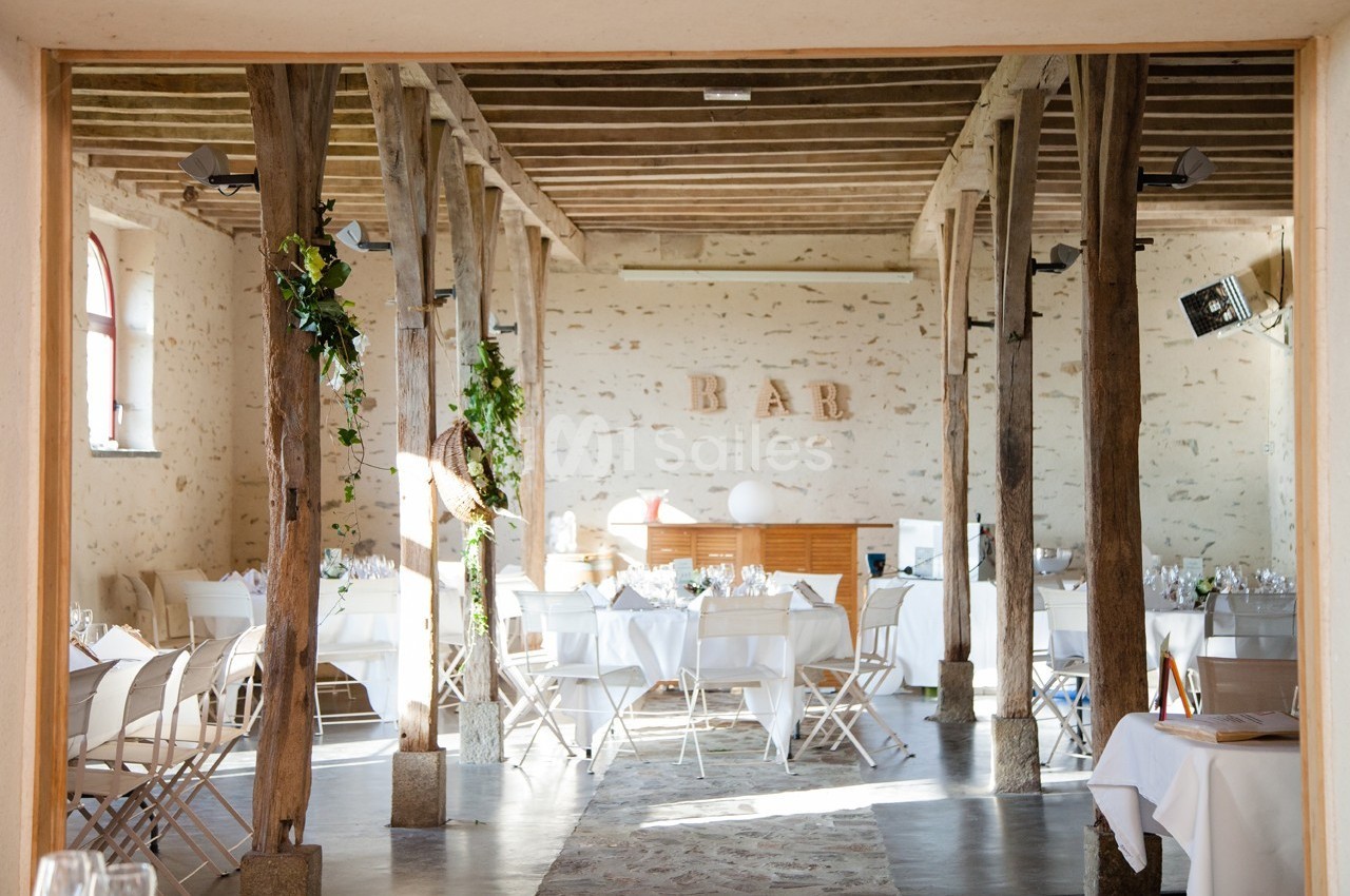 Salle de réception lumineuse avec poutres apparentes, tables dressées et espace bar en arrière-plan.