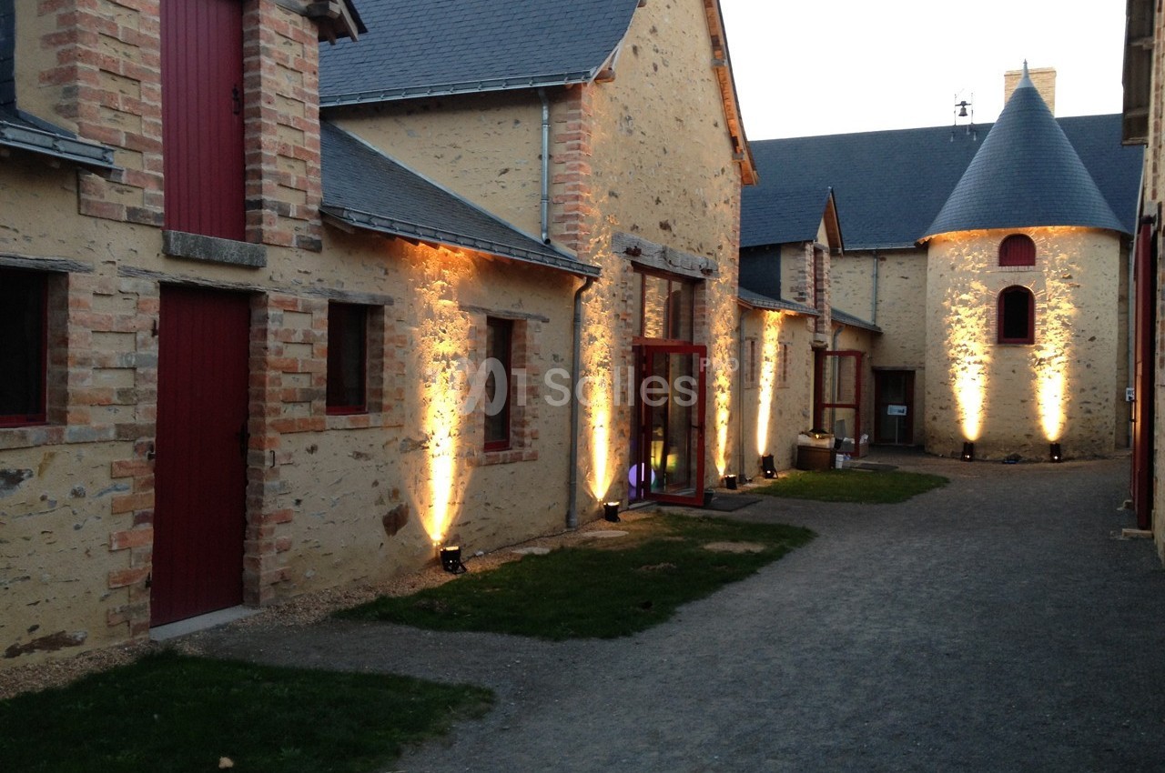 Bâtiments en pierre avec portes rouges, éclairés par des lumières au sol, dans une cour pavée au crépuscule.