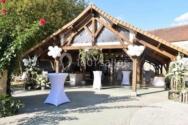 Structure en bois décorée pour un événement, avec ballons blancs, fleurs et tables hautes sous un ciel dégagé.