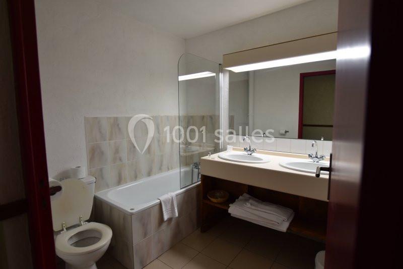 Salle de bain avec baignoire, double vasque, miroir éclairé, toilettes et serviettes pliées sur une étagère en bois.