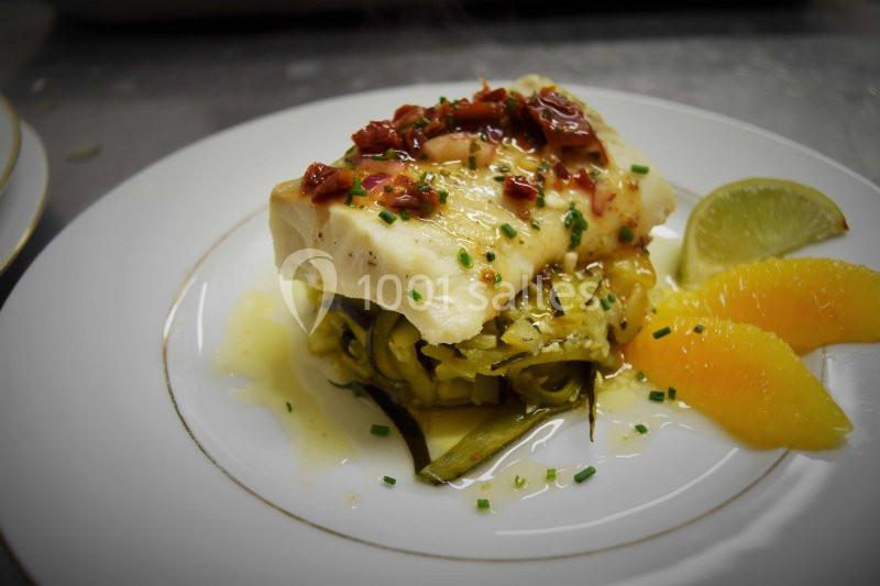 Filet de poisson servi sur un lit de légumes, garni de tomates séchées, herbes fraîches, et accompagné d'agrumes.