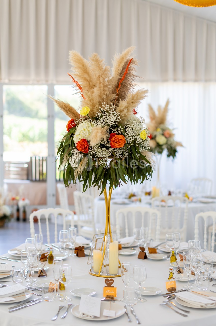 Centre de table floral avec pampas, roses orange et blanches, sur une table dressée dans une salle lumineuse.