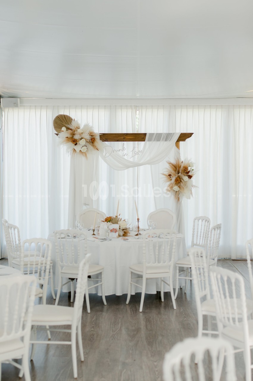 Table de réception décorée de fleurs séchées et voilage blanc, entourée de chaises blanches dans une salle lumineuse.