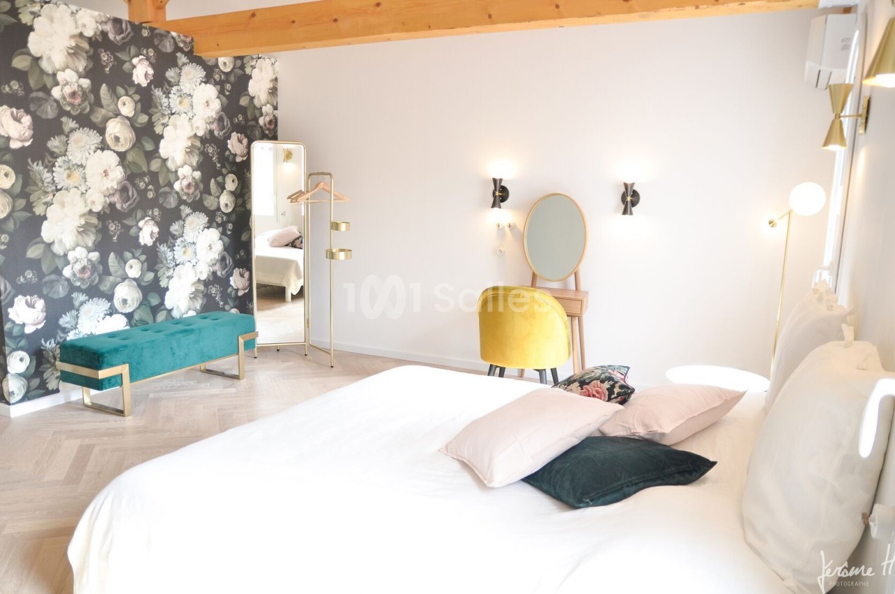 Chambre lumineuse avec lit double, mur fleuri, banc vert, coiffeuse jaune et poutres apparentes en bois.