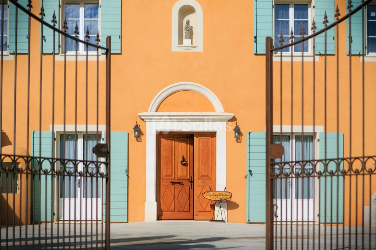 Façade d'une maison orange avec volets verts, porte en bois, statue en niche et portail métallique au premier plan.