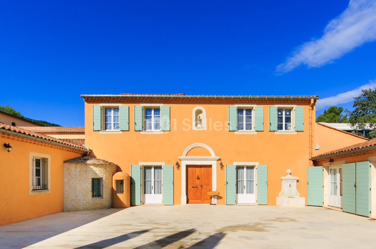 Façade d'une maison méditerranéenne avec murs orange, volets verts et cour ensoleillée sous un ciel bleu.
