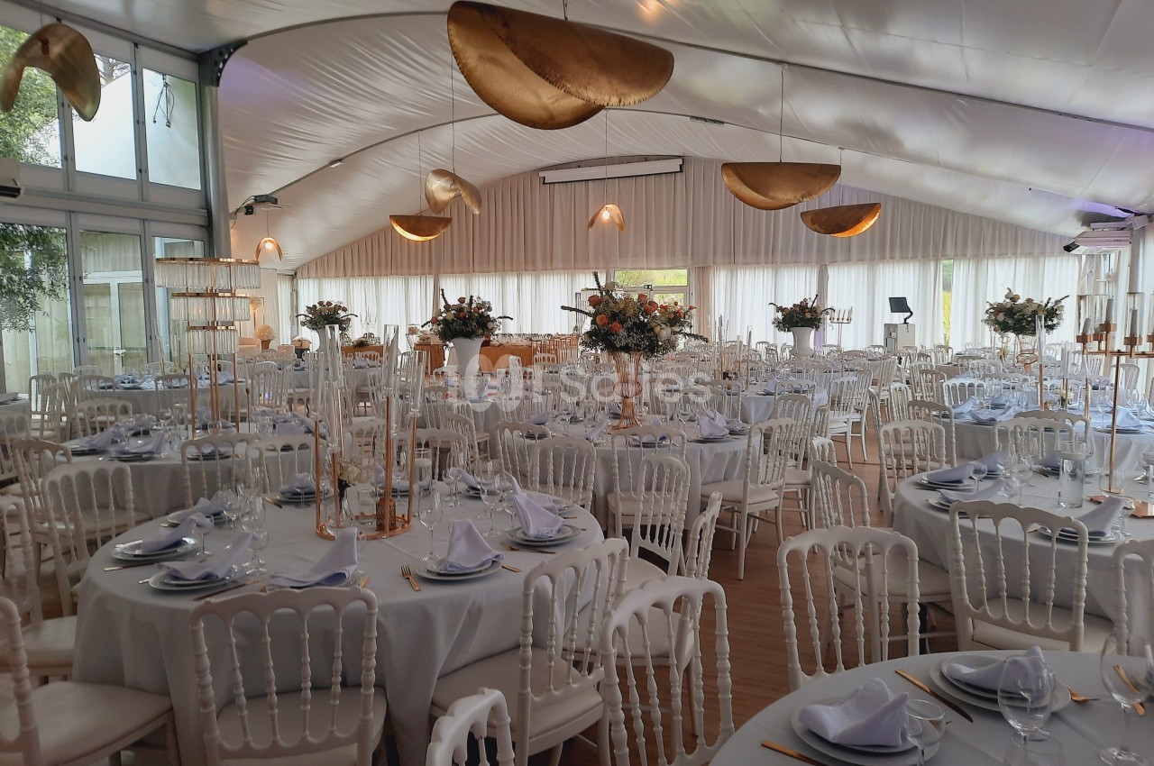 Salle de réception décorée avec des tables rondes dressées, nappes blanches, chaises élégantes et luminaires dorés.