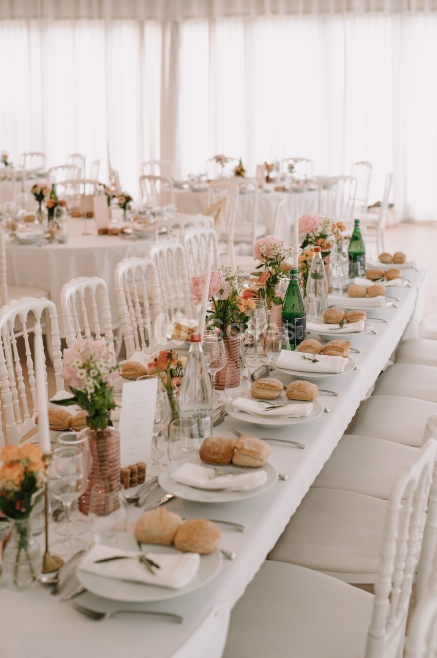 Table de réception élégamment dressée avec nappes blanches, fleurs, bouteilles d'eau et vaisselle disposée pour un repas.