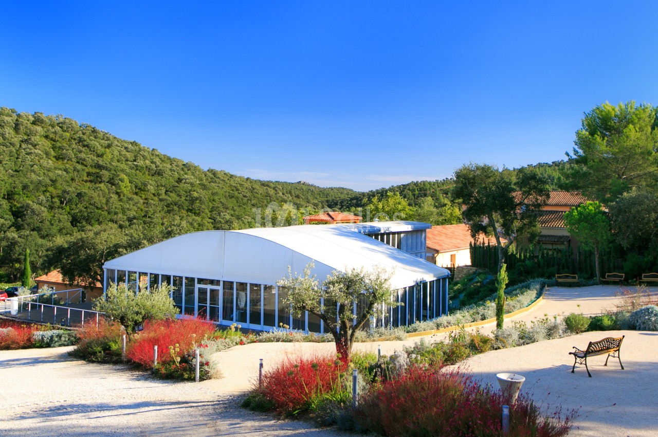 Vue d'un bâtiment moderne avec une structure blanche entouré de végétation méditerranéenne et d'un ciel bleu clair.