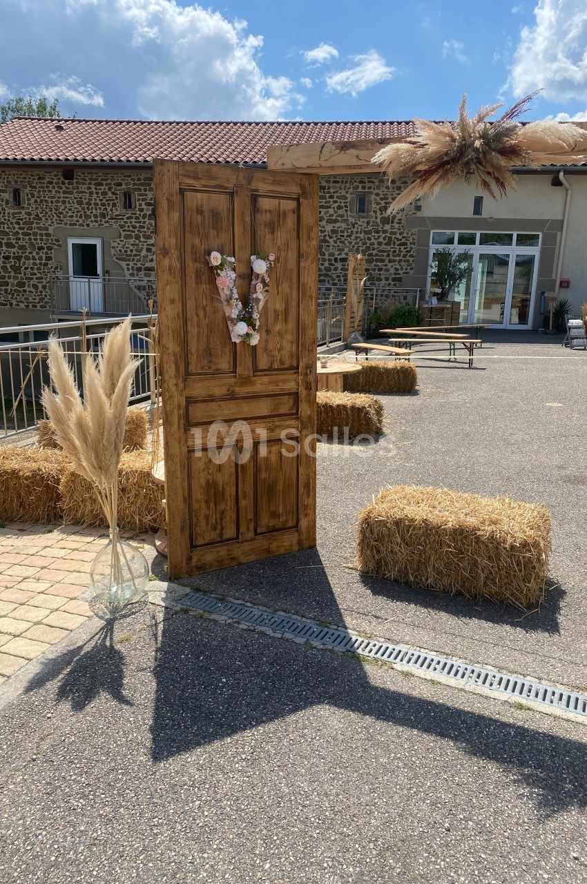Porte en bois décorée de fleurs, entourée de bottes de paille et de pampas, dans une cour extérieure ensoleillée.