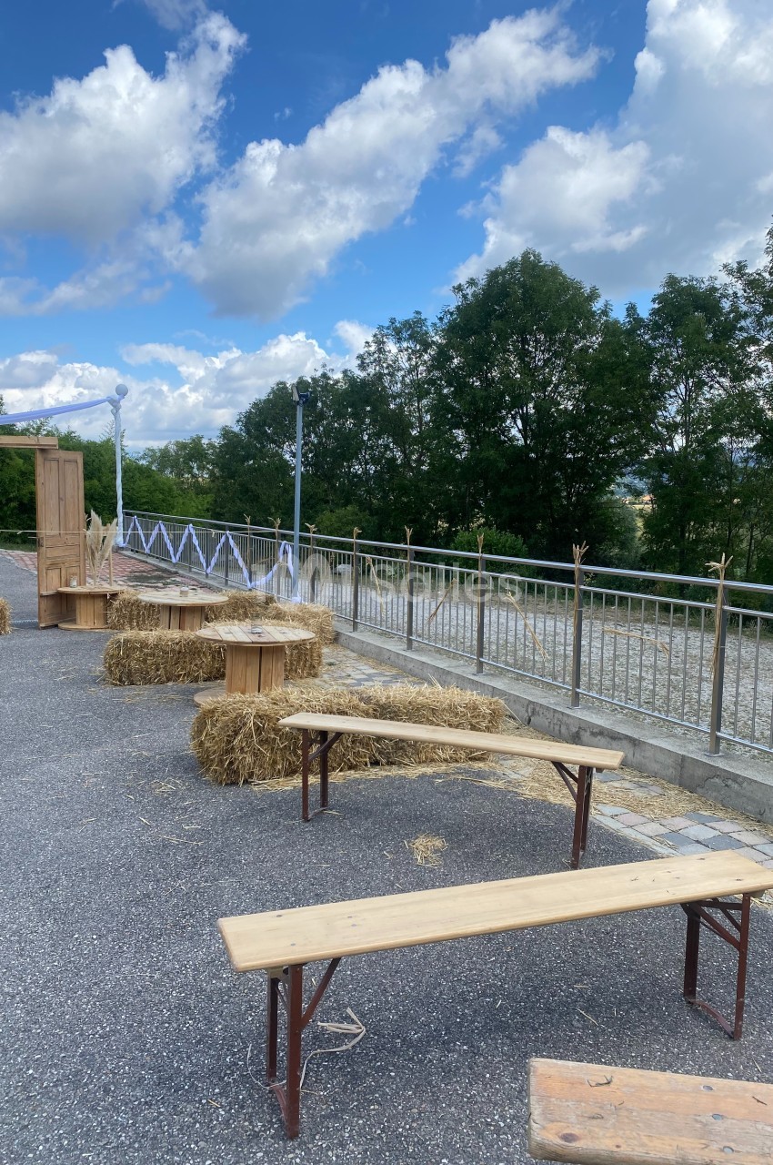 Bancs en bois et bottes de paille disposés en extérieur sur un sol pavé, avec des arbres et un ciel nuageux en arrière-plan.